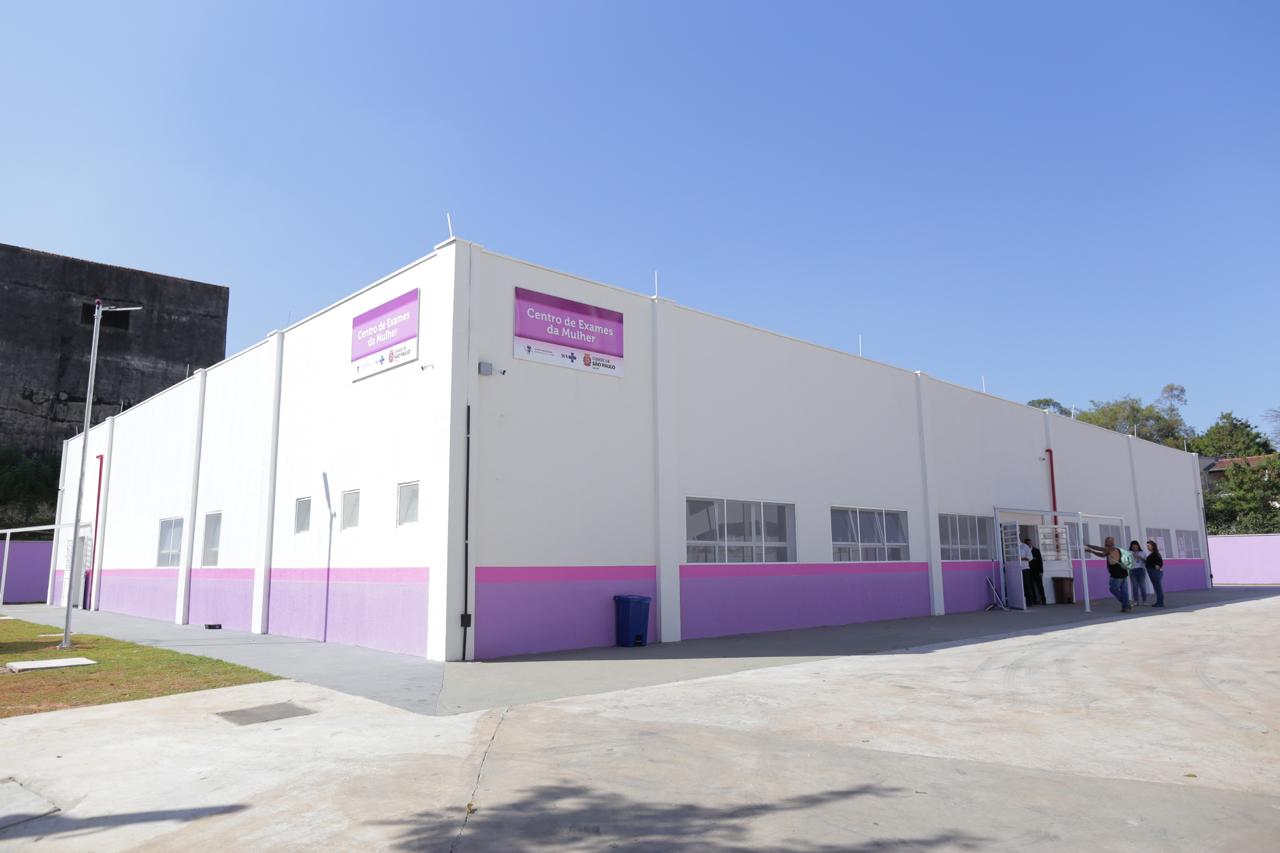 A imagem mostra um prédio branco com uma faixa rosa e uma roxa na base. O prédio tem uma placa onde é possível ler "Centro de Exames da Mulher". 